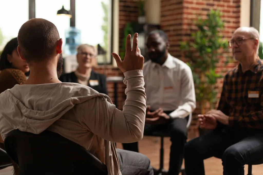 A group of people sits in a circle indoors, engaged in discussion. One person, seen from behind, raises a hand while others listen attentively. The setting appears casual and supportive, with warm lighting and plants.